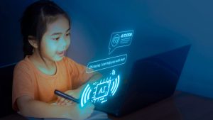 A young girl sits at a desk in a dark room, focused on her laptop. She writes in a notebook while chatting with an AI tutor displayed on the screen.