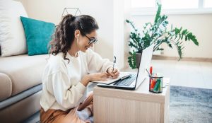 aucasian woman is doing online lessons at the laptop smiling while writing something sitting on the floor
