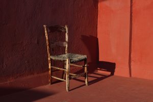 Old wooden one vintage chair in a room with red grunge walls and floor in sunlight, dark shadows. Shabby antique furniture. Empty copy space, no people. Retro interior still life, concept.