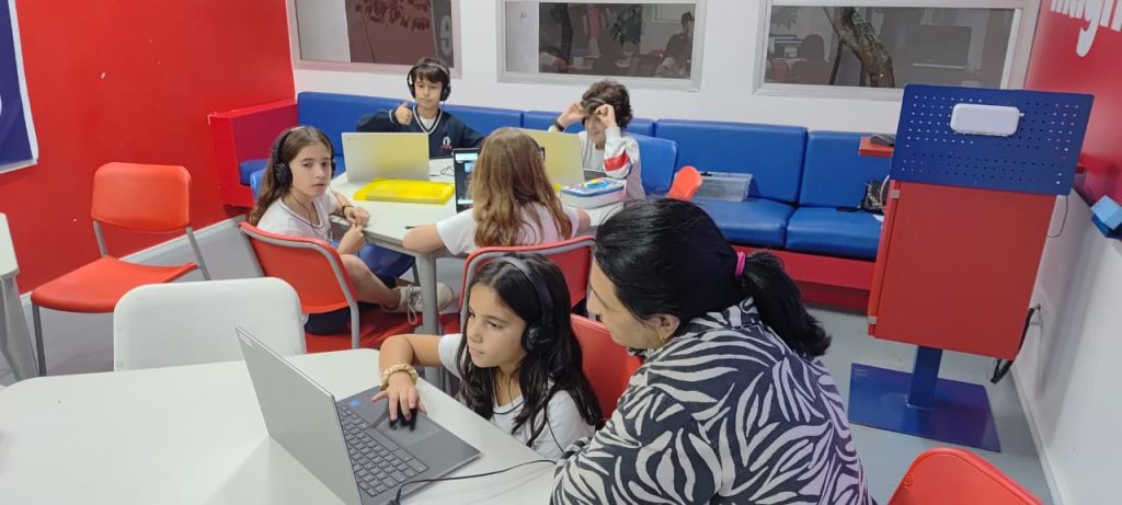 Student listening to a laptop while a teacher looks on. Other students using laptops in the background.
